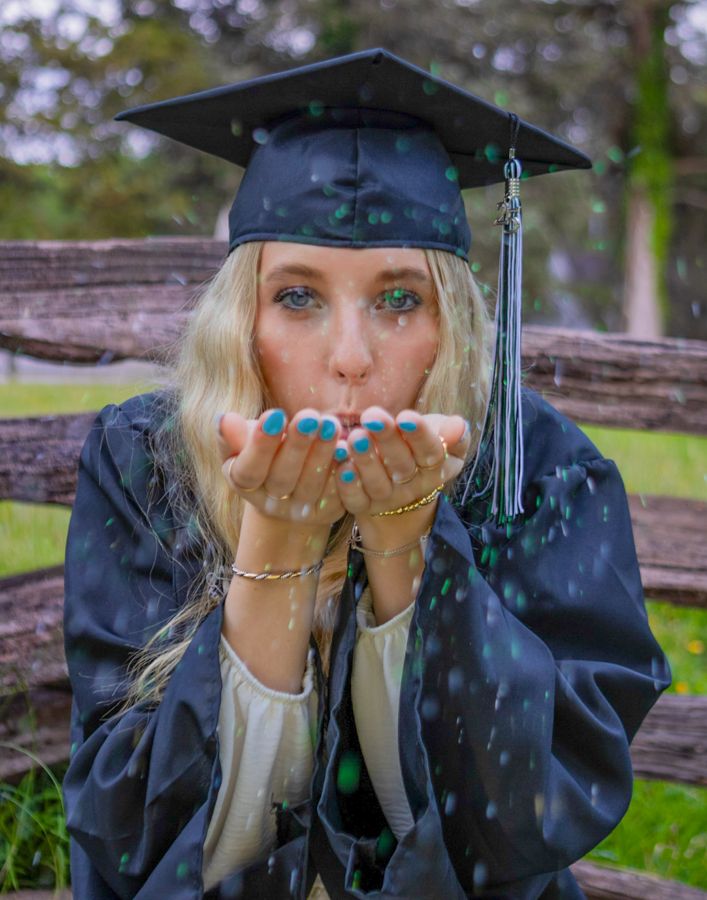 Blonde graduate in cap and gown blowing glitter from hands outdoors.