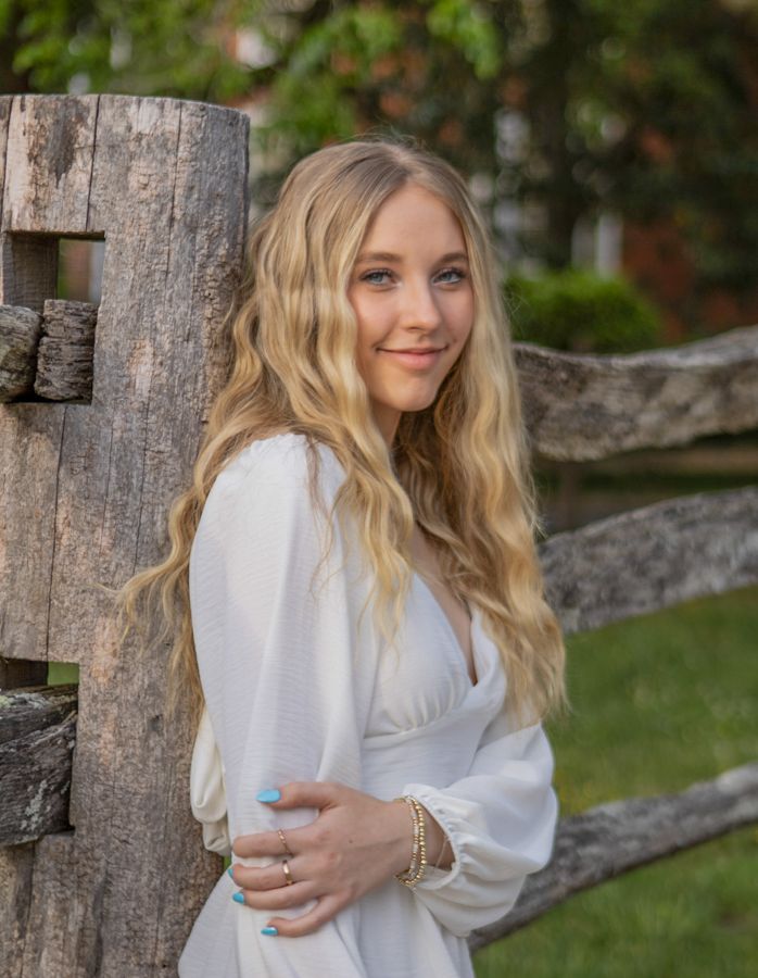 Beautiful blonde teenage girl in a white dress leaning against a rustic wooden fence for a senior portrait session.