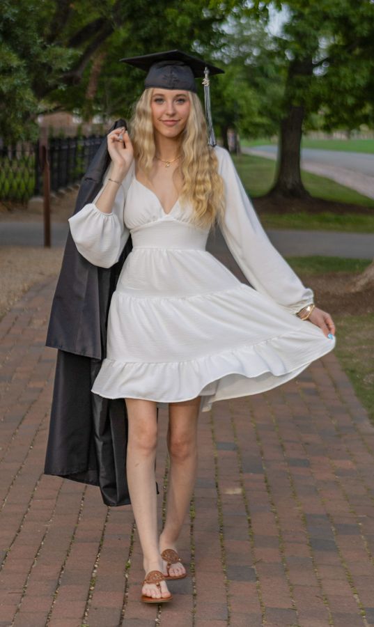 Young blonde woman in white dress and graduation cap holding black gown on a tree-lined brick pathway.