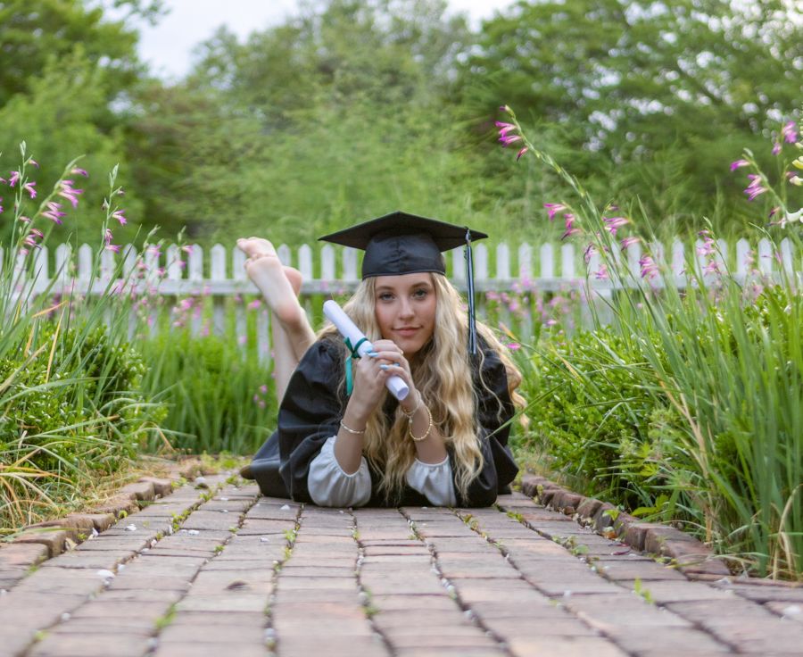 Smiling graduate lying on brick path holding diploma in lush garden.