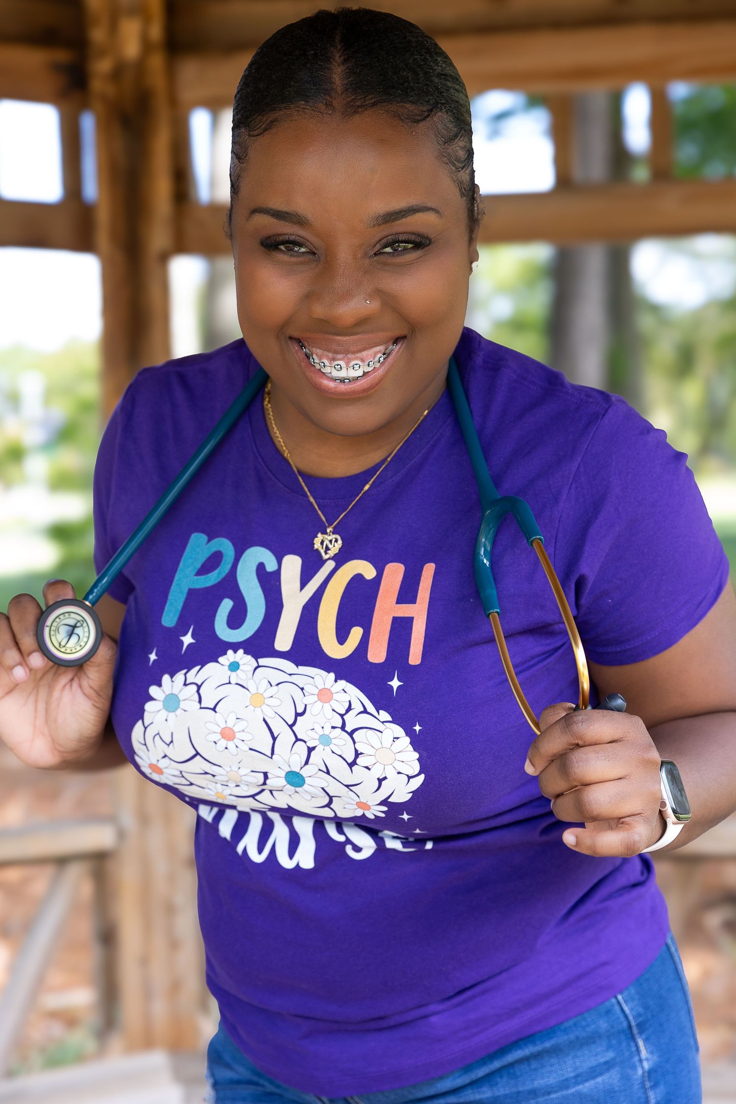 Smiling woman wearing a purple Psych Nurse t-shirt with a stethoscope around her neck outdoors.
