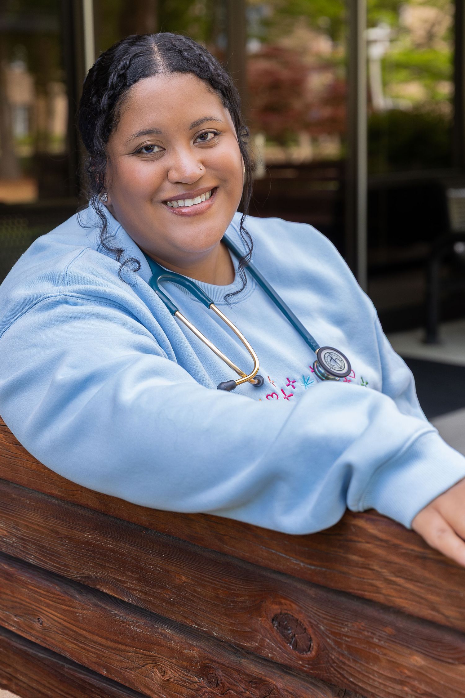 Smiling female nurse in blue scrubs with stethoscope sitting at outdoor wooden table.