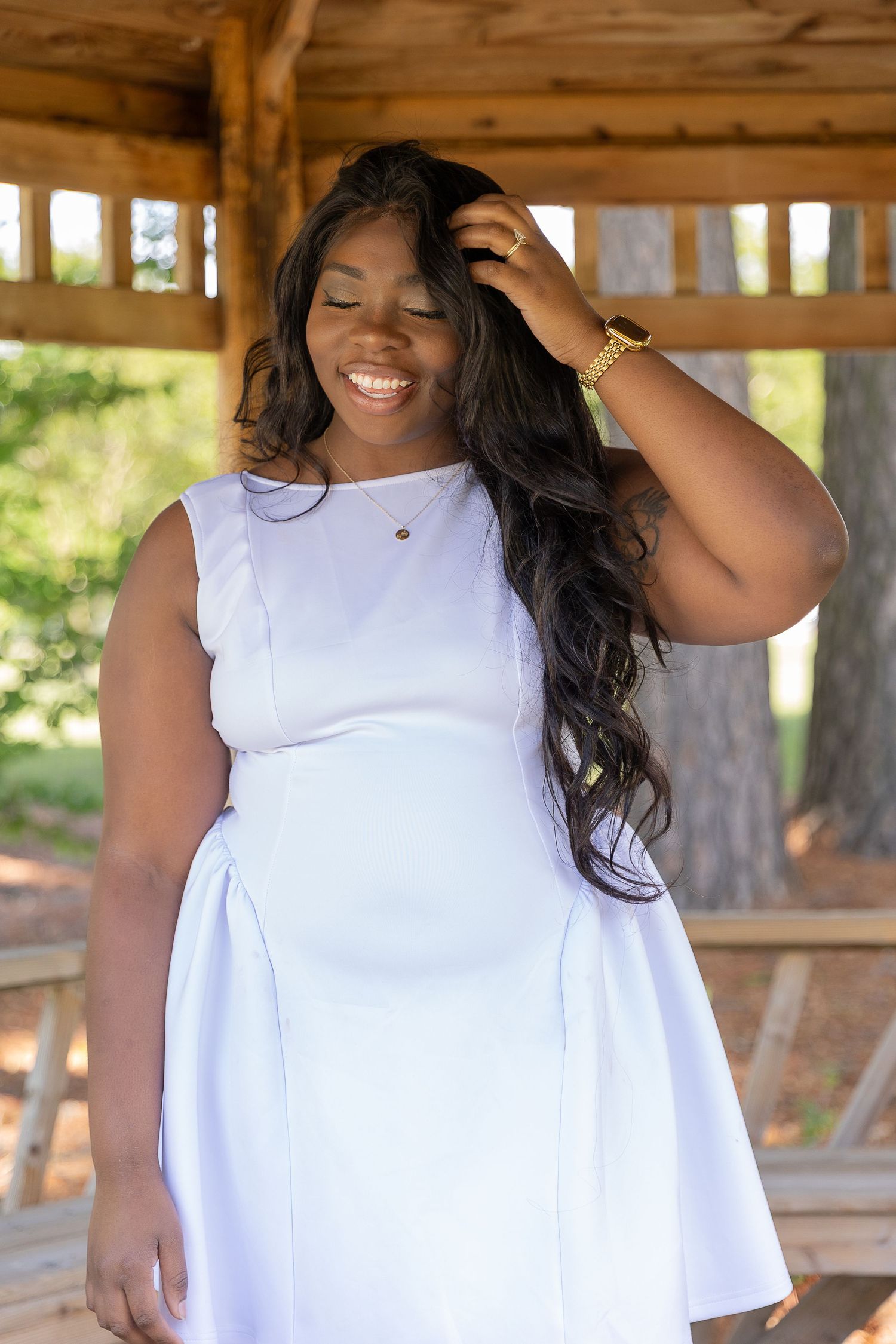 Smiling young Black woman in white sleeveless dress poses outdoors under wooden gazebo with long wavy hair.