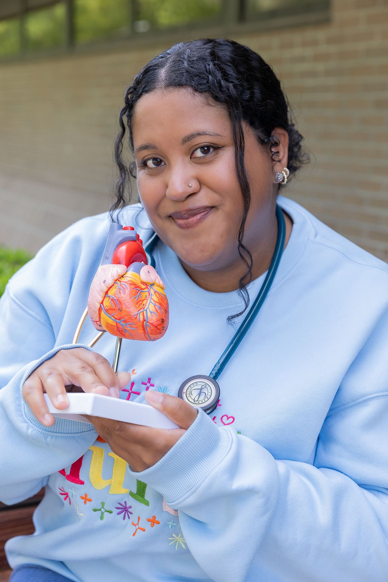 Female nursing student holds anatomical heart model wearing stethoscope and light blue medical sweatshirt.