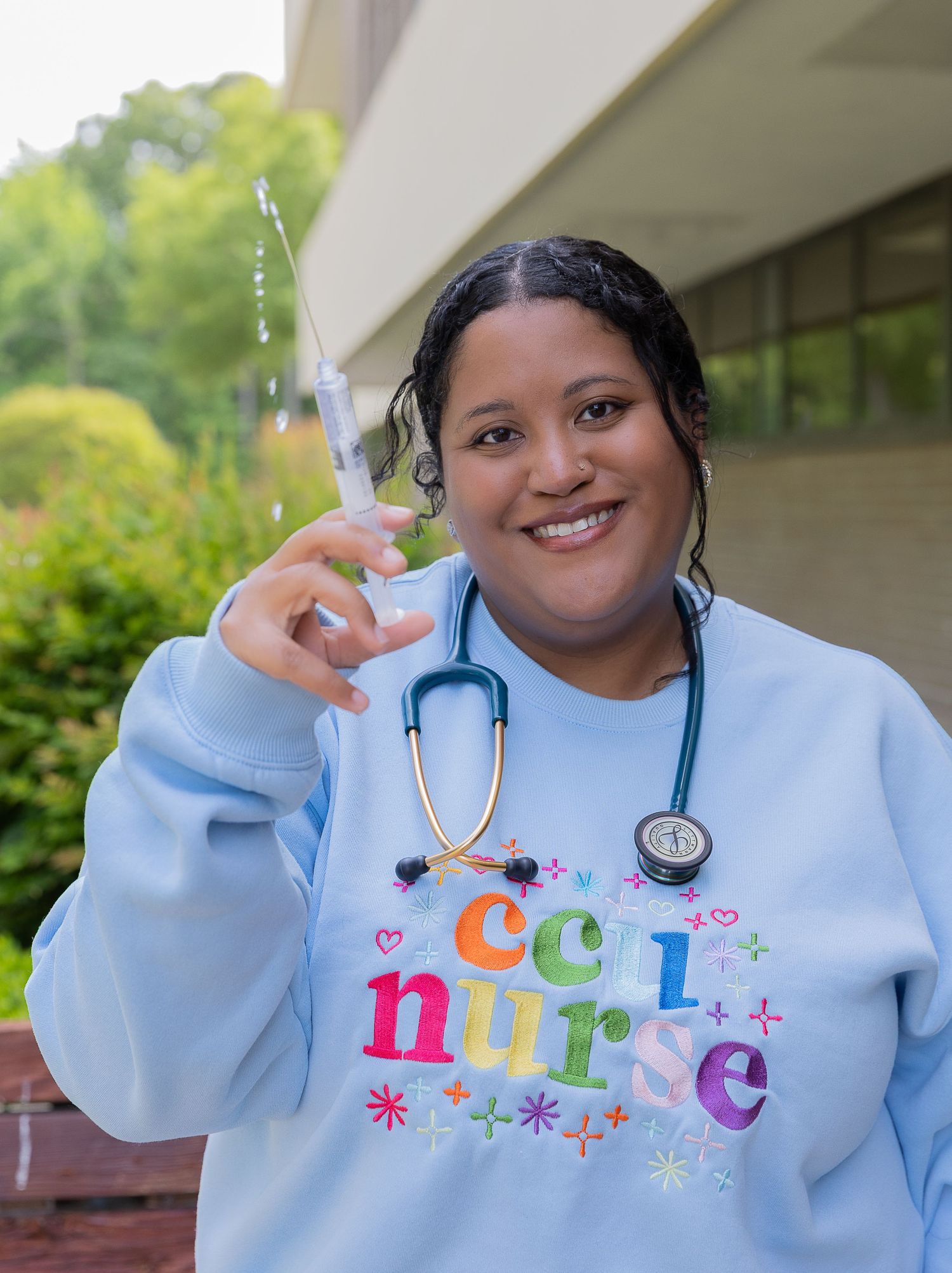 Smiling CCU nurse wearing a light blue sweatshirt holds a syringe with a teal stethoscope around her neck.