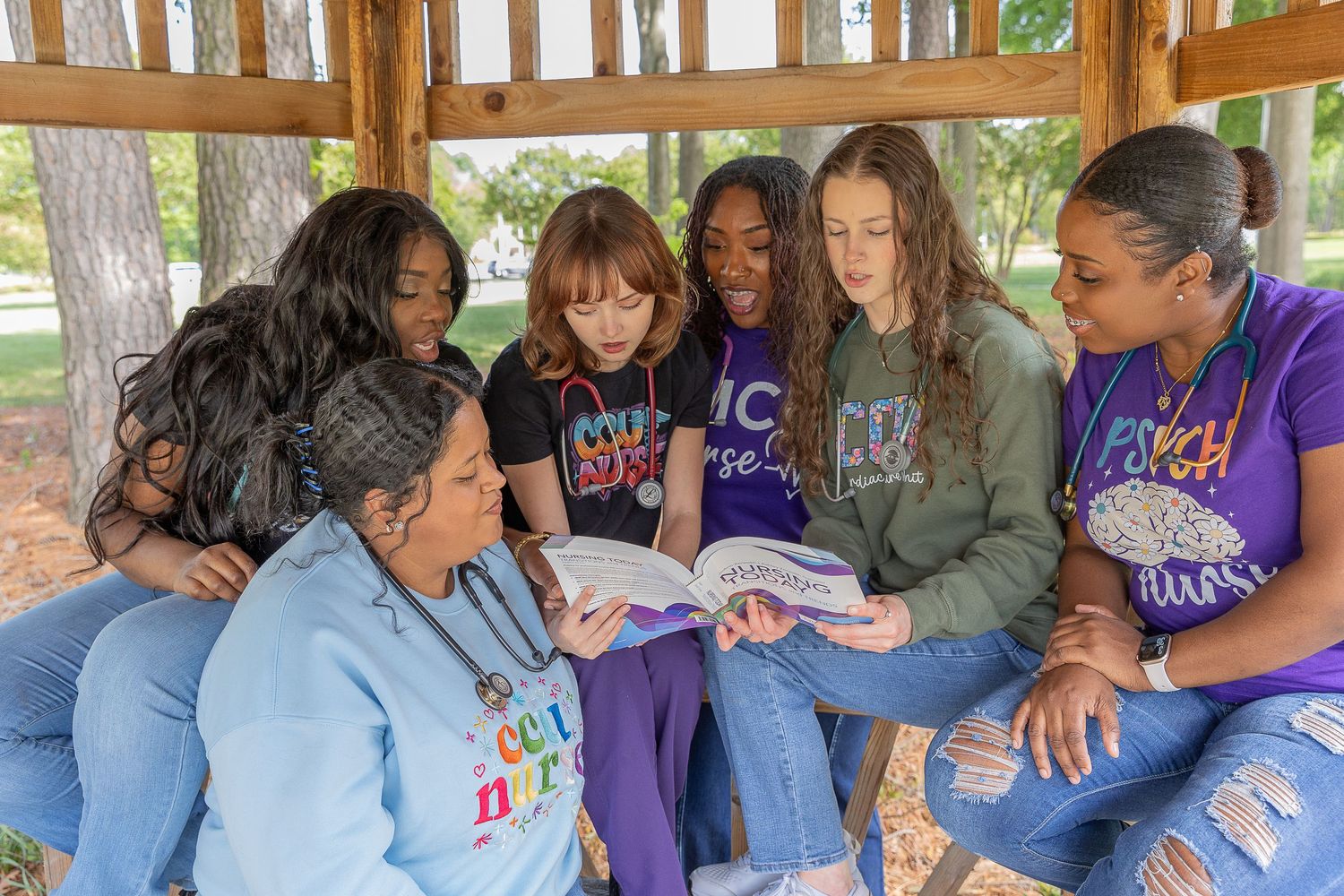 Six girls sitting under a gazebo reading a book together outdoors in a park setting.