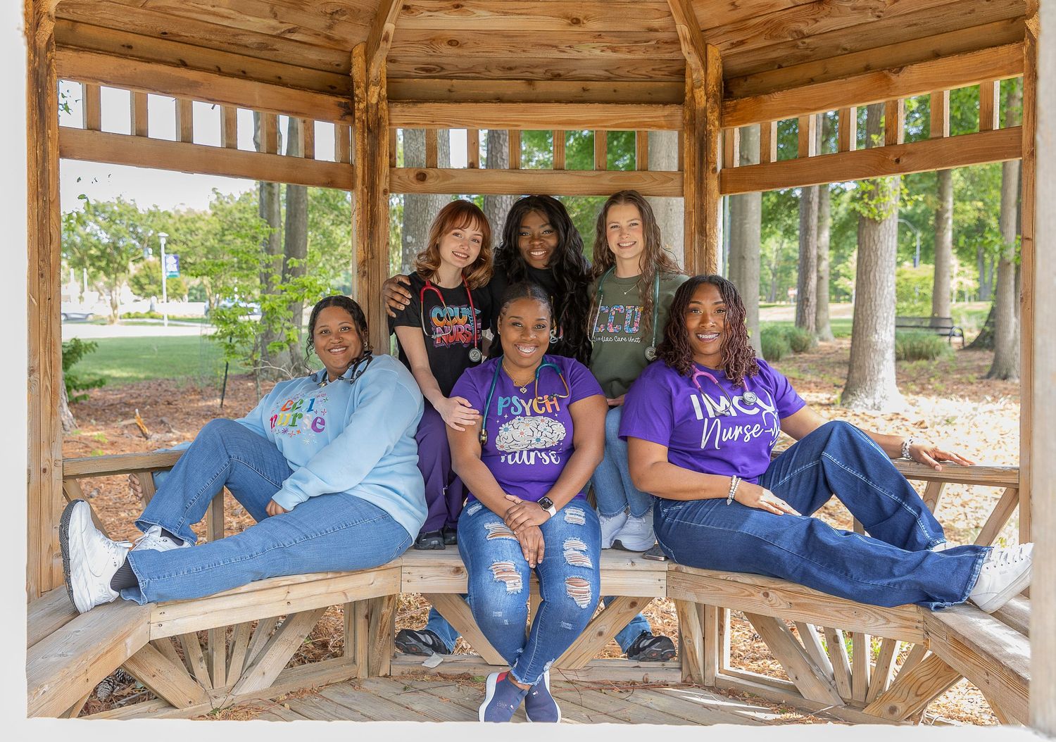 Six women smiling together under a wooden gazebo in a park, wearing casual and college-themed clothing.