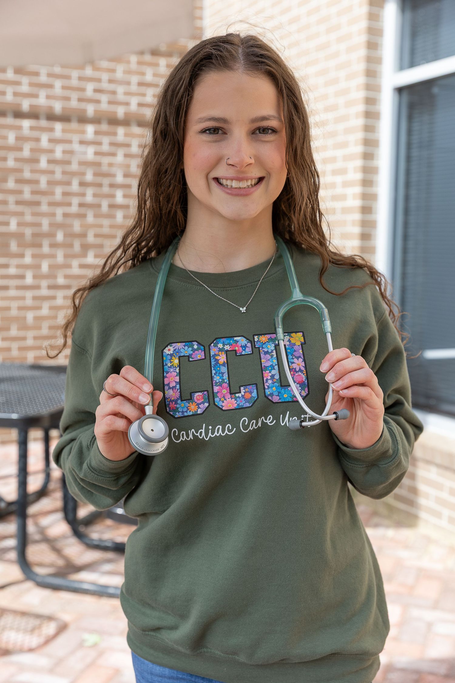 Smiling young woman in olive CCU Cardiac Care Unit sweatshirt holds a stethoscope outdoors.