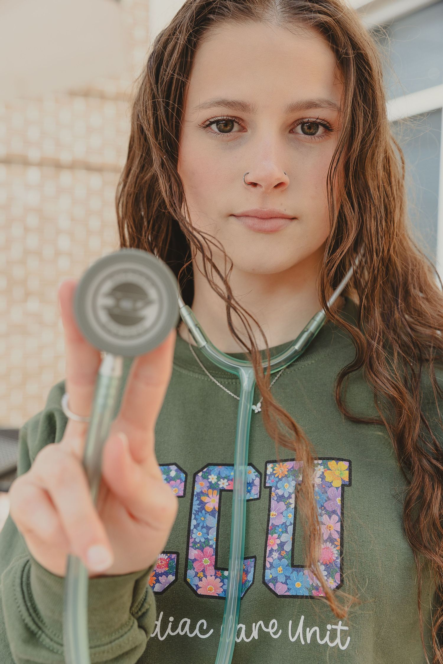 Young girl in olive CCU sweatshirt holds stethoscope toward camera with serious expression.