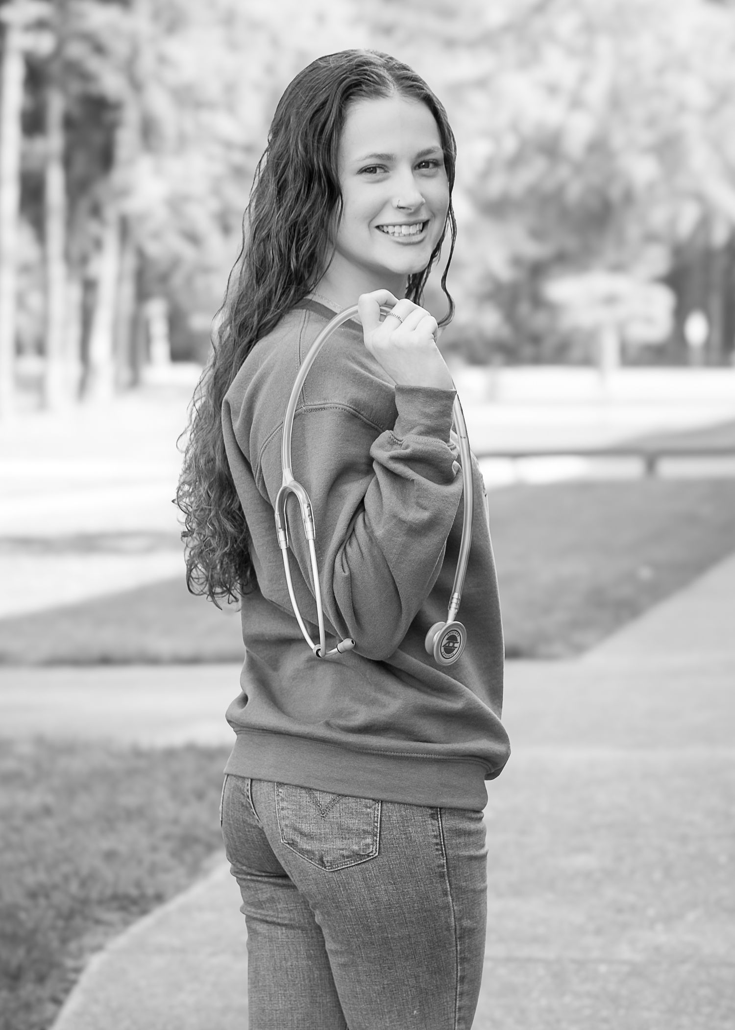 Smiling young woman with curly hair holding a stethoscope over her shoulder in a park setting.