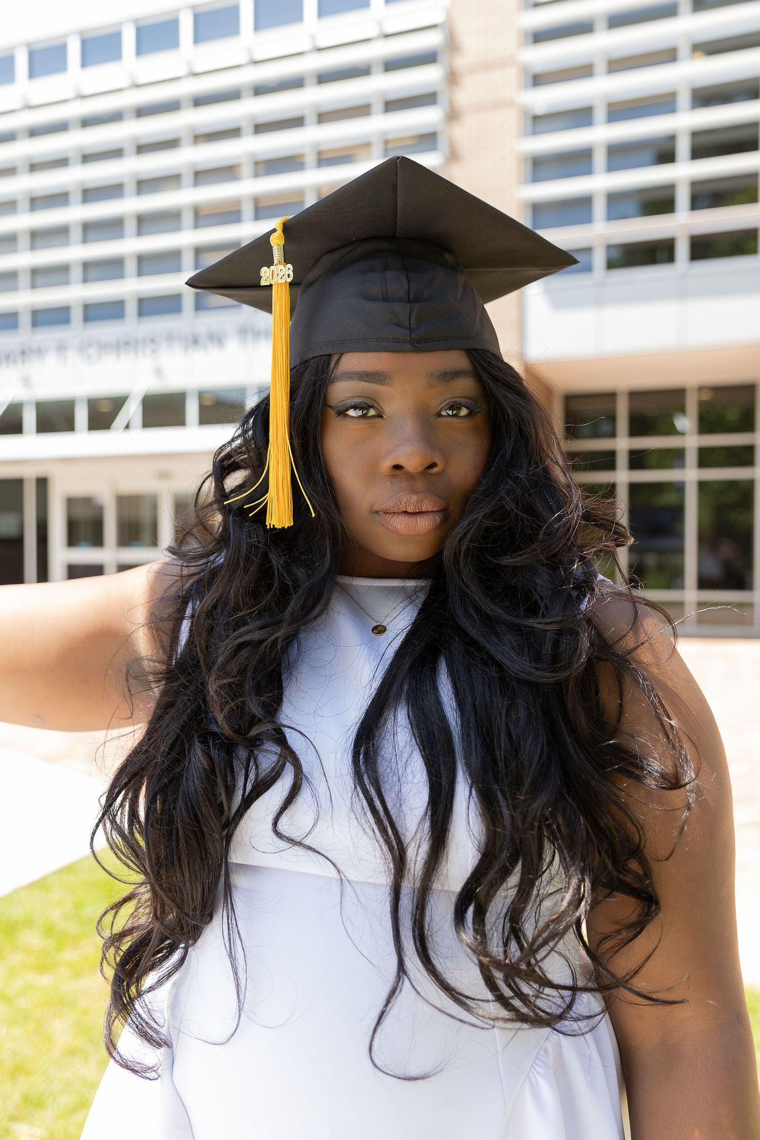 Young Black woman in graduation cap with gold tassel and white dress poses outdoors near university building.