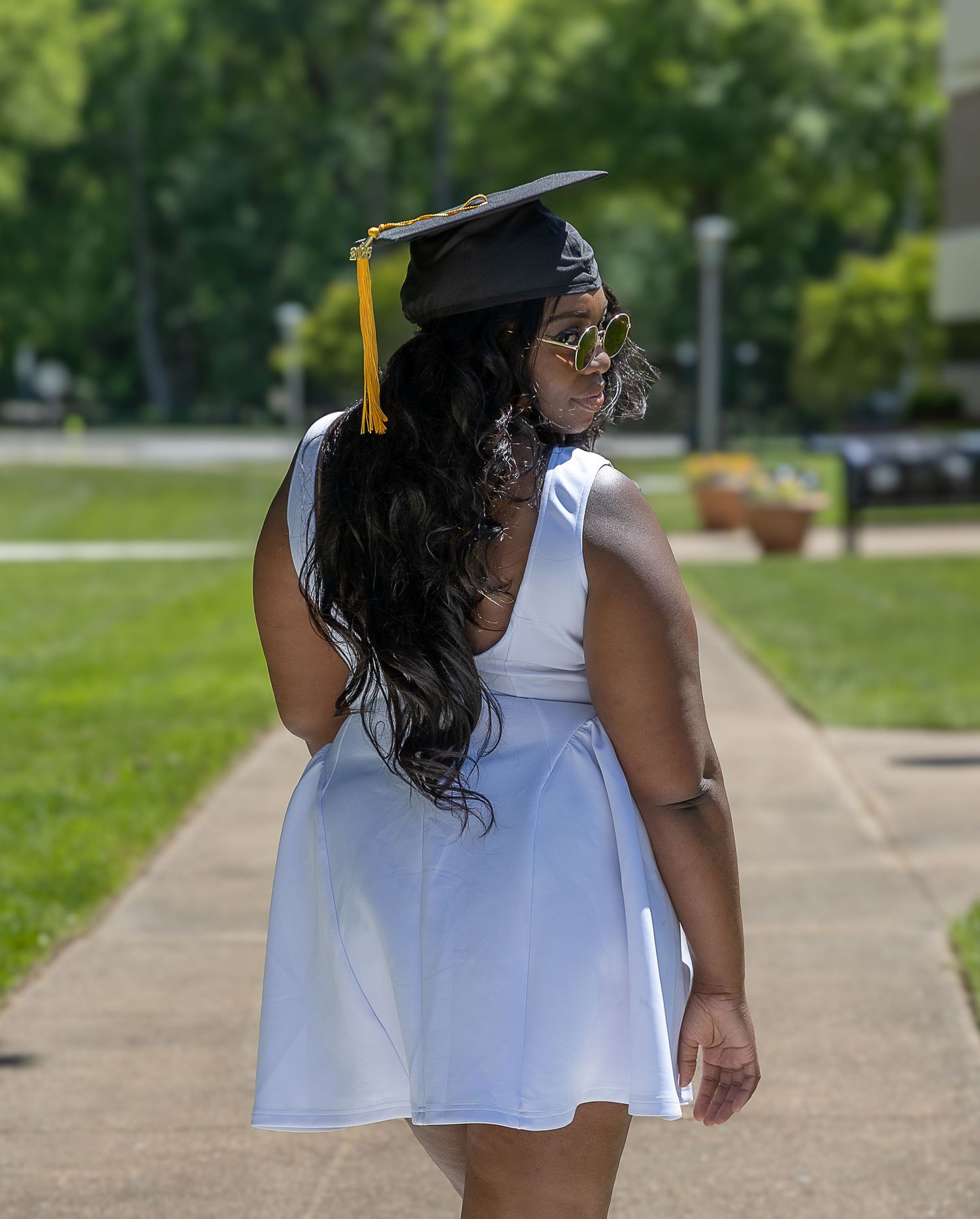Young woman in white dress and graduation cap with gold tassel poses on a sunny campus walkway.