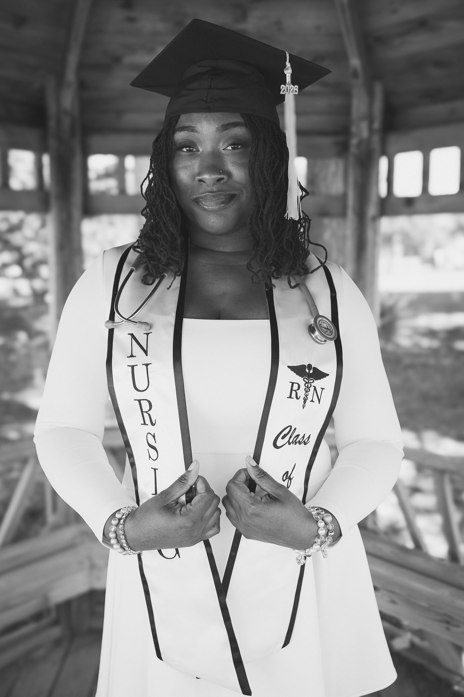 Black and white photo of a nursing graduate wearing cap, gown, and RN Class stole, posing proudly under a pavilion.