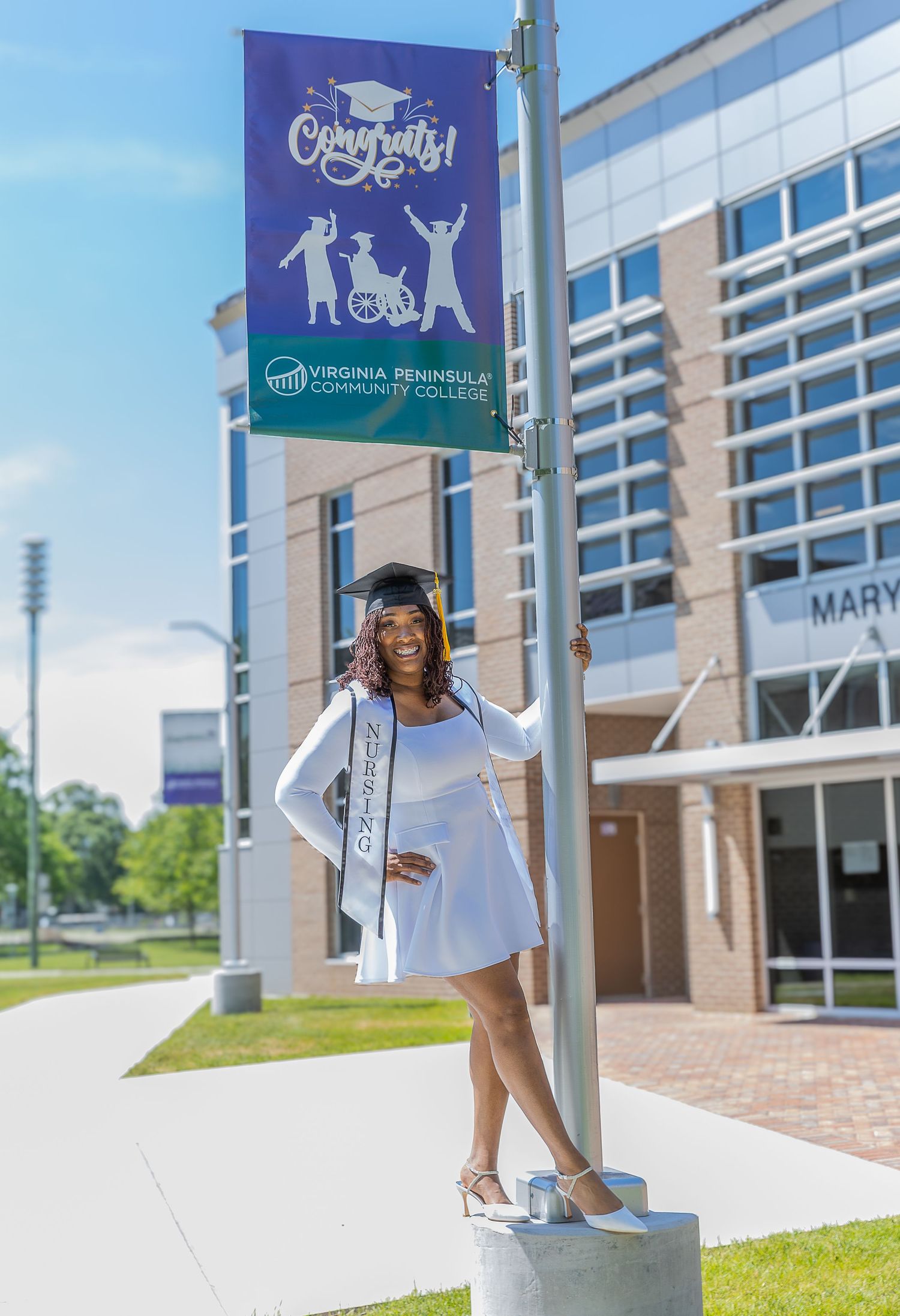 Graduate in white dress and cap poses by Virginia Peninsula Community College Congrats banner on sunny day.