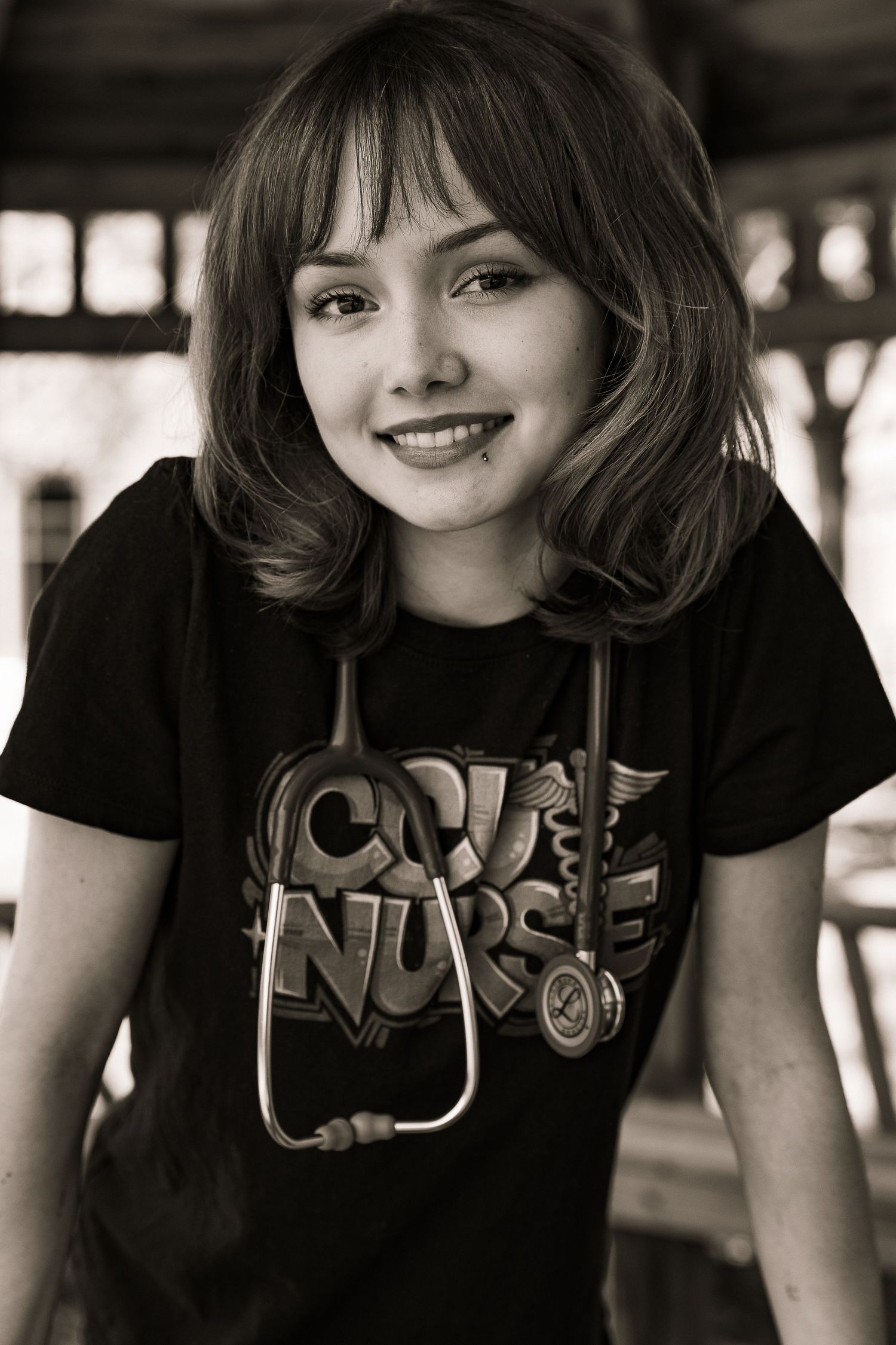 Smiling young woman wearing a black nurse t-shirt with stethoscope around her neck in sepia tone.