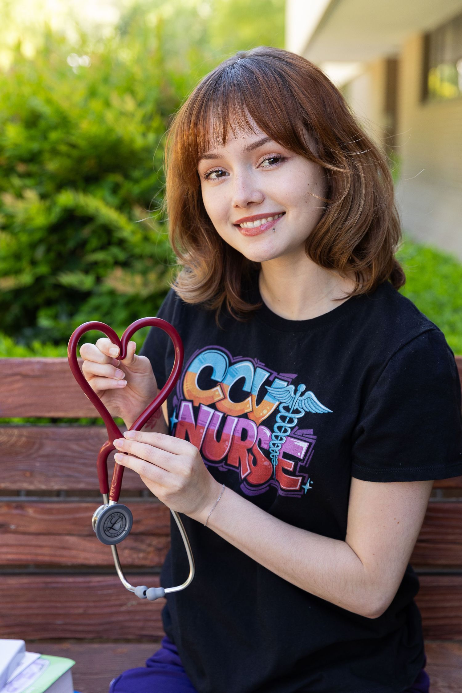 Young nursing student in CCU Nurse t-shirt holds red stethoscope shaped as heart, smiling outdoors on bench.
