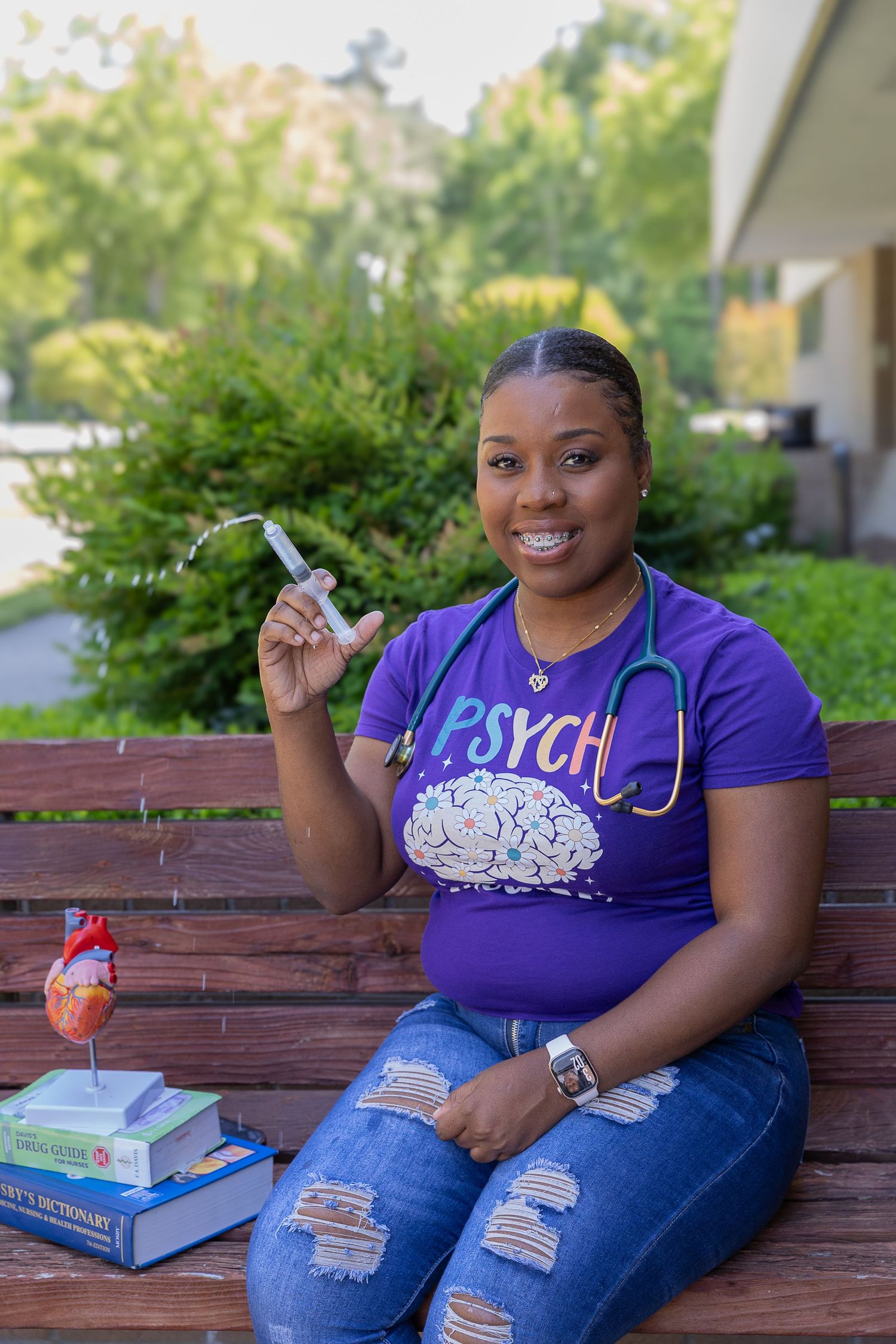 Smiling nursing student in purple PSYCH shirt holding syringe, wearing stethoscope, seated on bench with medical books.