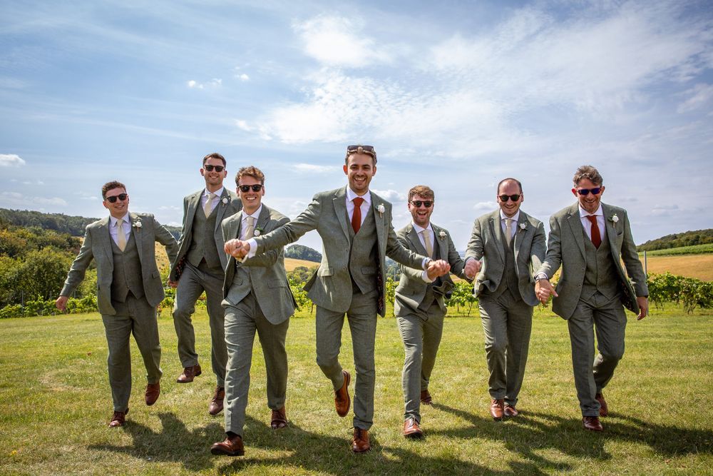 Groomsmen in matching grey suits walk confidently across sunny vineyard field on wedding day.