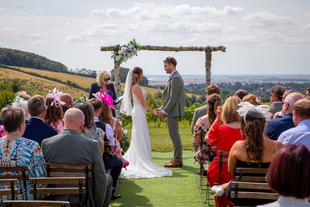 Couple stand at outdoor altar beneath wooden arch with breathtaking rolling countryside landscape behind them.