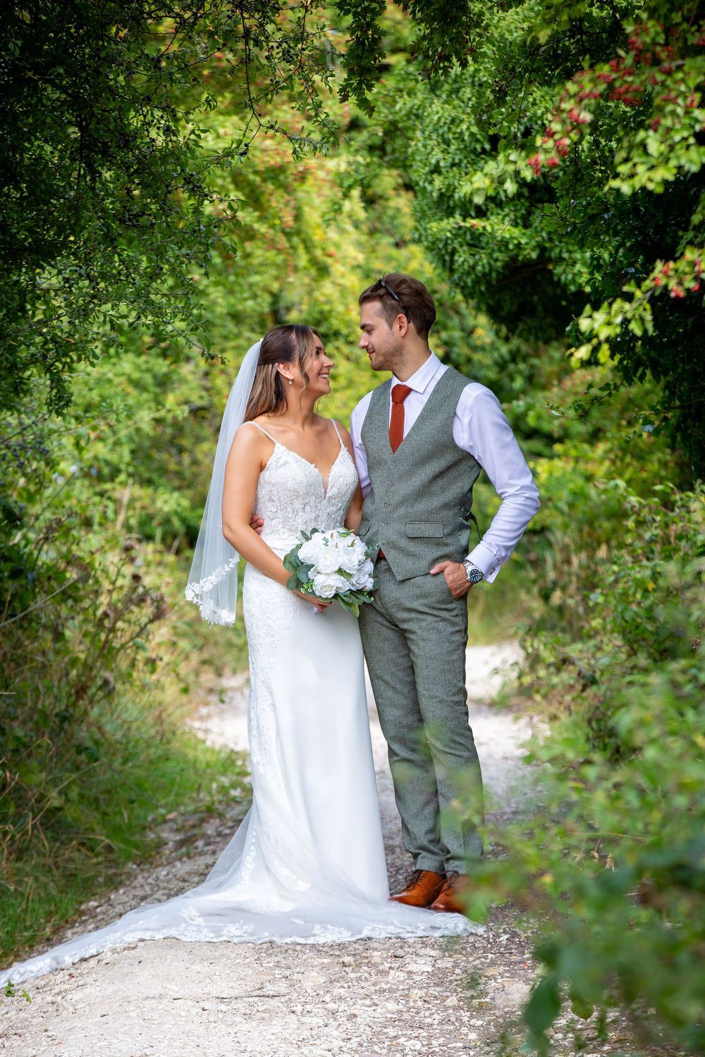 Bride and groom share romantic moment on tree-lined path, bride holding white bouquet in lace gown.