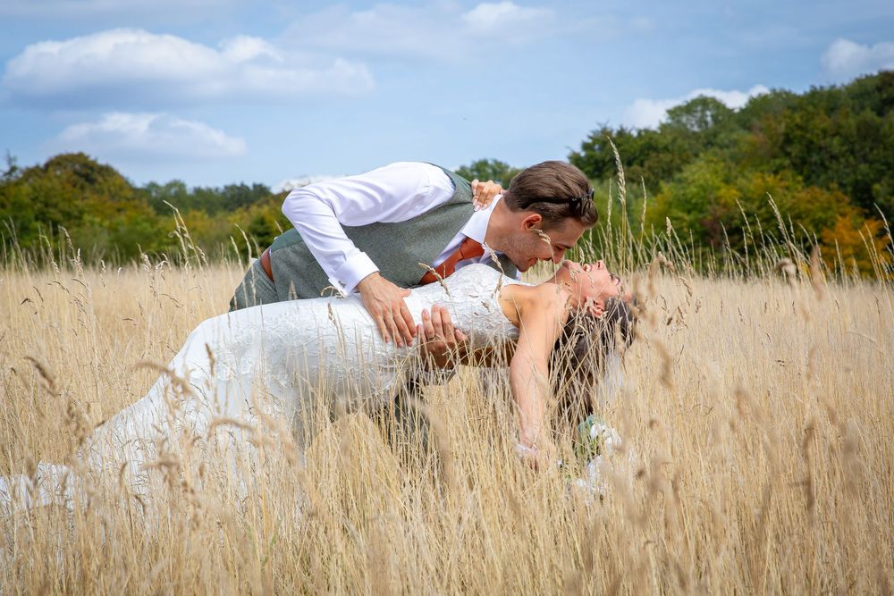 Groom dips bride romantically in golden wheat field under blue sky on wedding day.