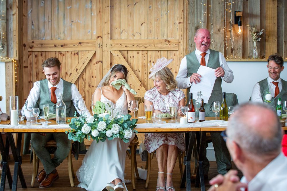 Bride and groom laugh during wedding speech at rustic barn reception with floral centerpiece.