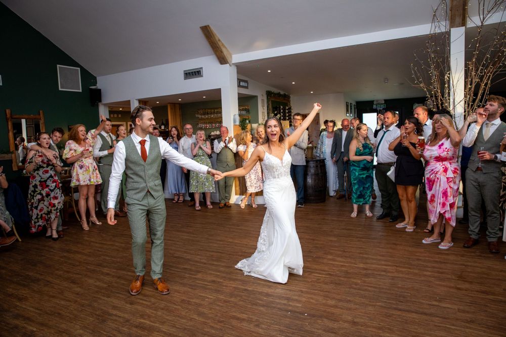 Bride and groom share first dance with arms raised before cheering wedding reception guests.