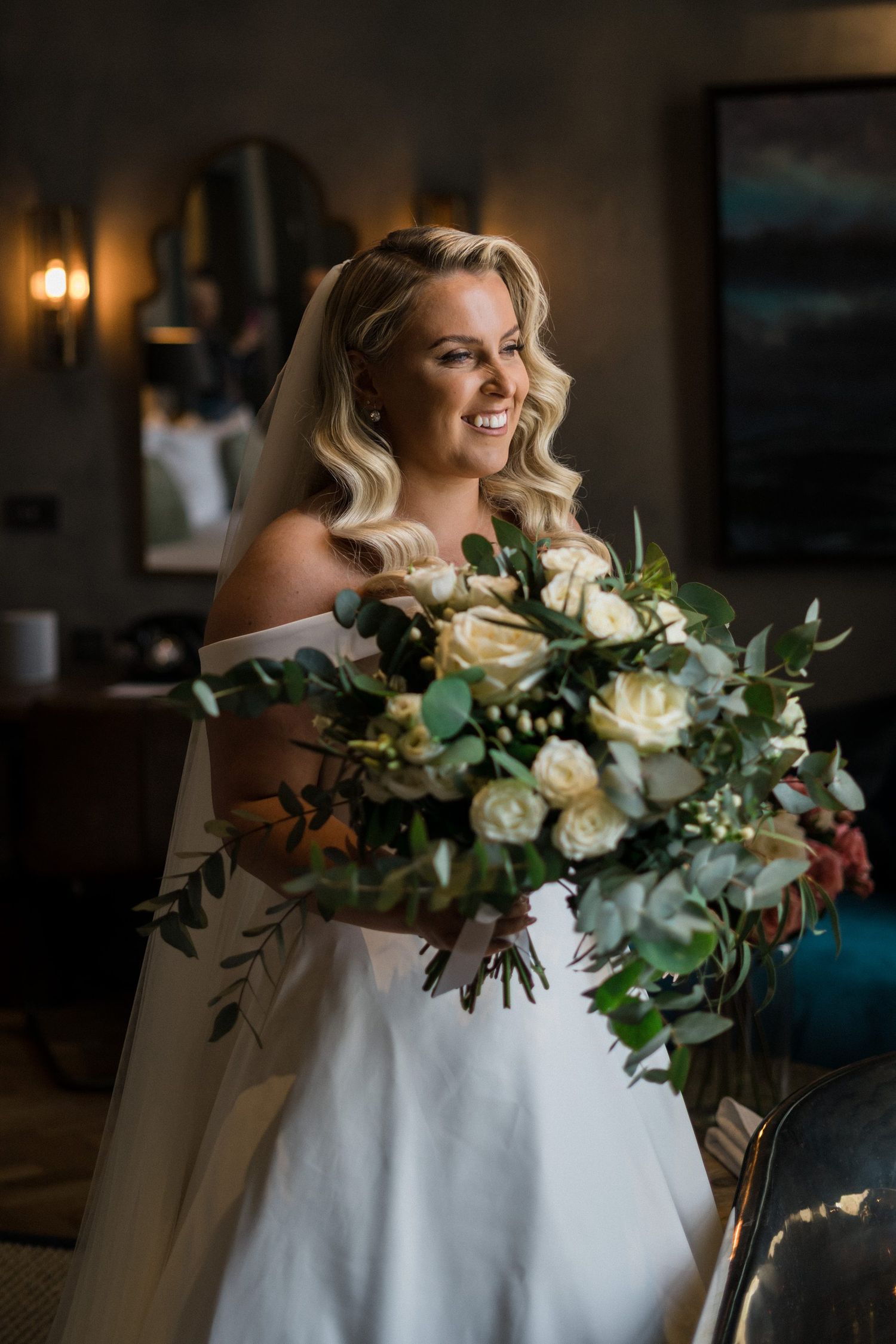 Elegant white wedding dress with a romantic off-shoulder design adorned with lush greenery and cream rose bouquet details.