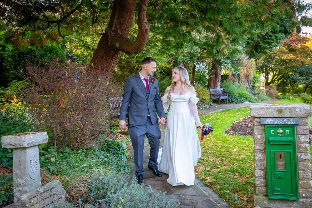 Newlywed couple walks hand in hand through colorful autumn garden past green letterbox.