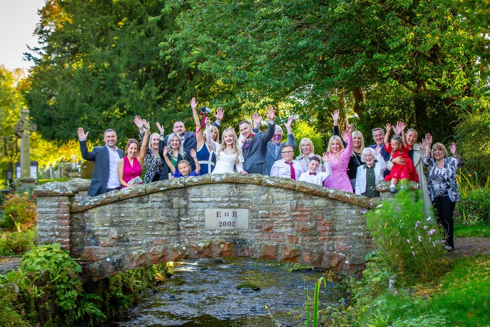 Wedding party celebrates on rustic stone bridge waving at camera surrounded by lush green trees.
