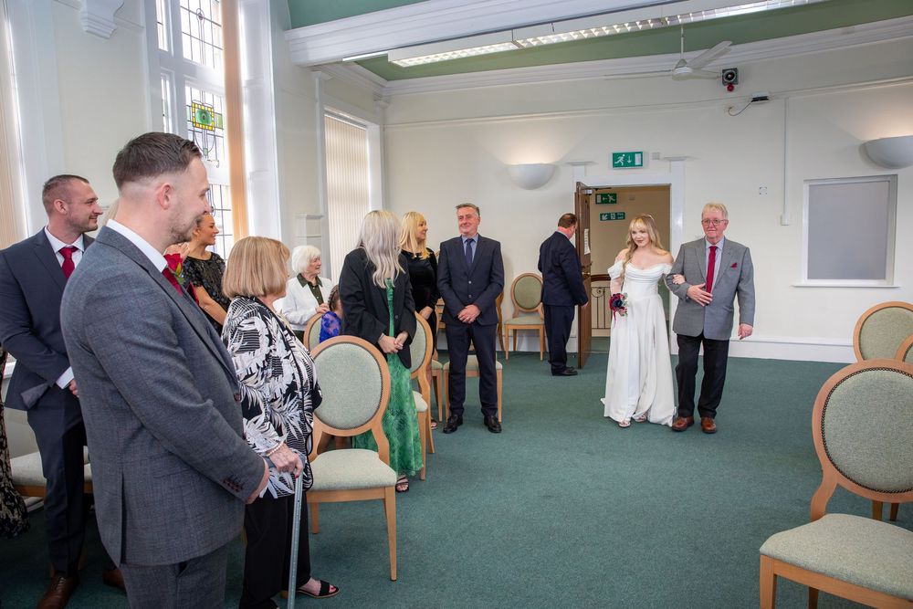 Bride walks down aisle in bright ceremony room while groom waits at the front.
