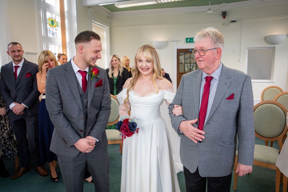 Bride in white gown walks down aisle with older man while groom in grey suit watches during wedding ceremony.