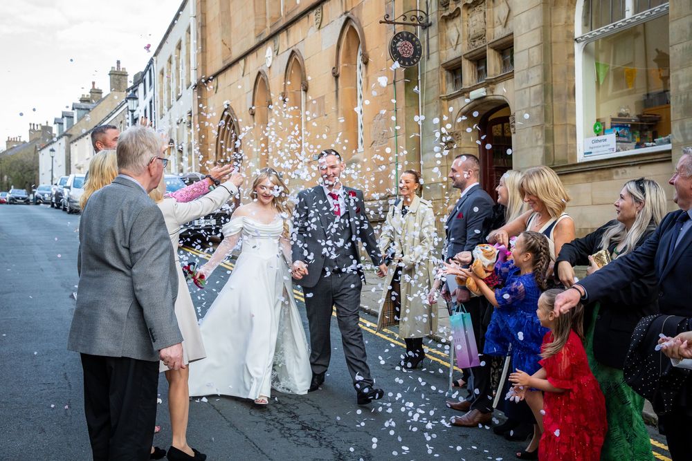 Joyful newlyweds walk through confetti shower surrounded by cheering guests outside historic stone building.