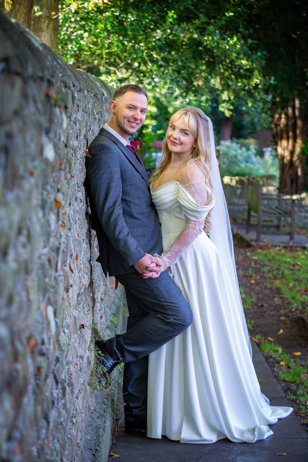 Smiling bride and groom pose together against rustic stone wall in tree-lined garden path.
