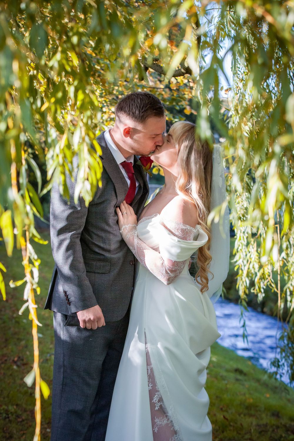 Newlyweds share tender kiss framed by weeping willow branches in sunlit outdoor setting.