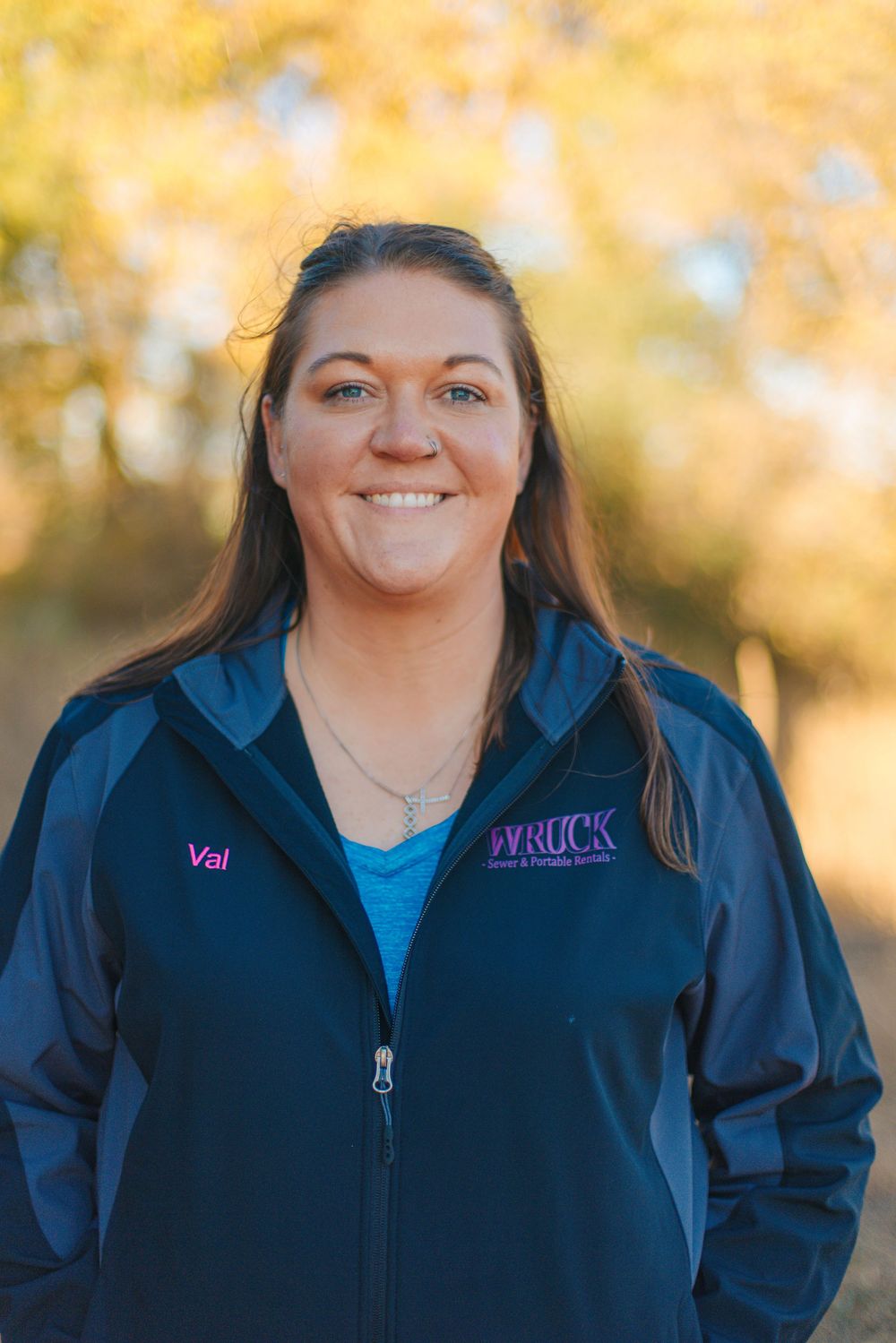 Woman named Val smiling outdoors in a navy Wruck jacket, with autumn foliage in the background.