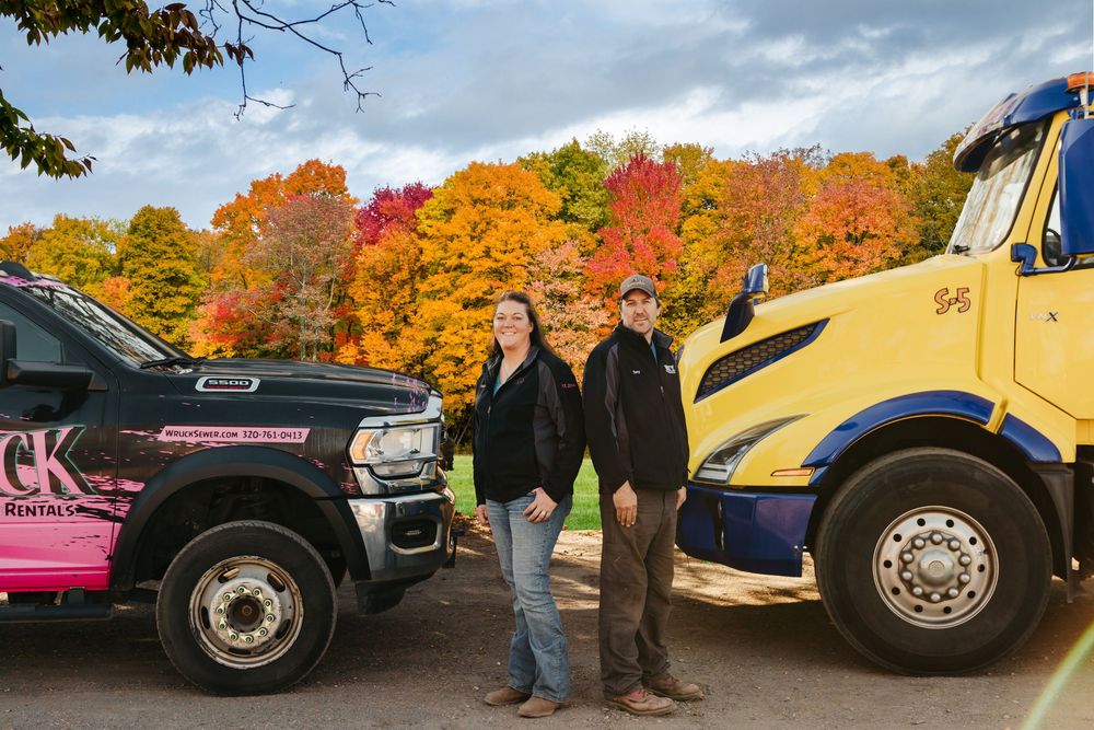 Two people stand between a black pickup truck and yellow semi-truck surrounded by vibrant fall foliage.