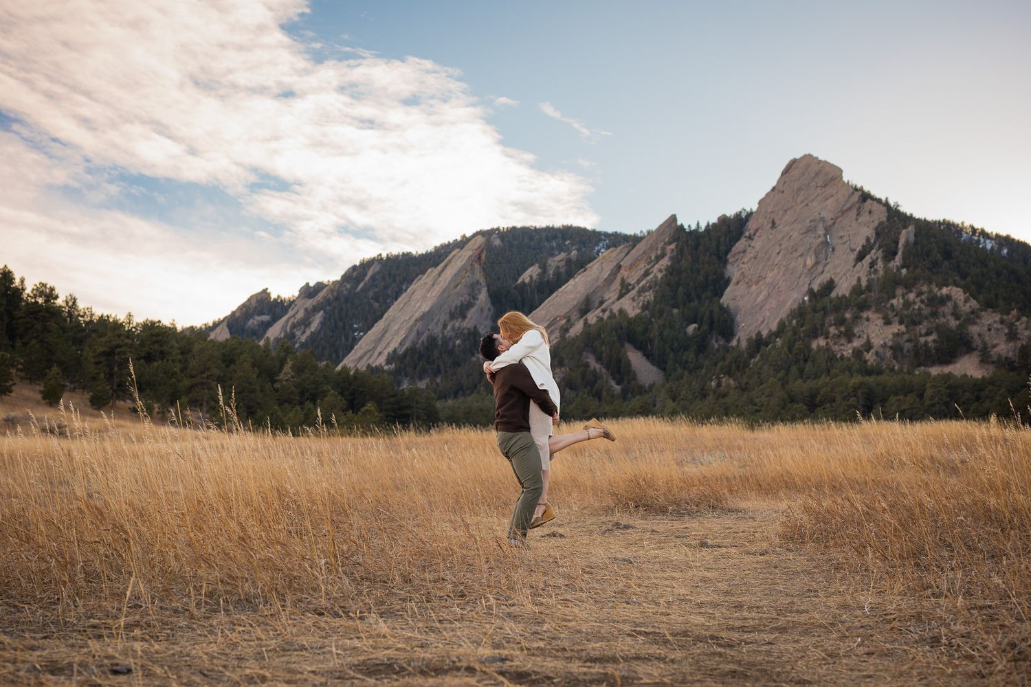 A person jumps joyfully in a golden meadow with the dramatic Flatirons rock formation rising in Boulder, Colorado.