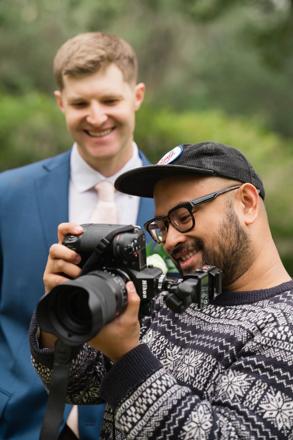 Professional photographer wearing a patterned sweater and baseball cap takes photos with a DSLR camera outdoors.