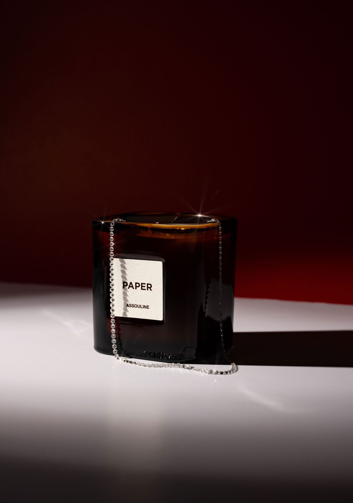 Assouline Paper scented candle in amber glass jar with silver chain, displayed against dark red background.