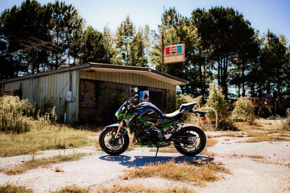 Green Kawasaki motorcycle parked outside an abandoned rural building with overgrown grass and pine trees.