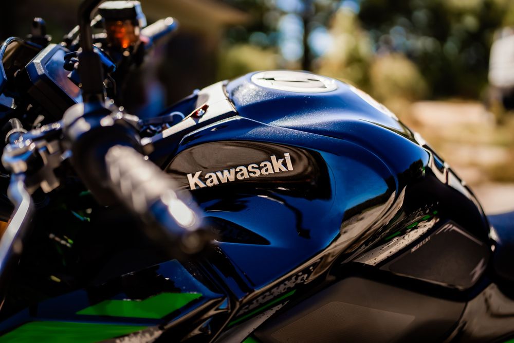 Close-up of a Kawasaki motorcycle fuel tank showcasing the brand logo, with green accents and bokeh background.