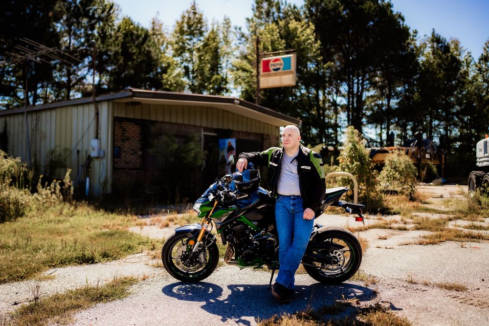 Motorcyclist posing beside a green Kawasaki bike at an abandoned roadside building on a sunny day.
