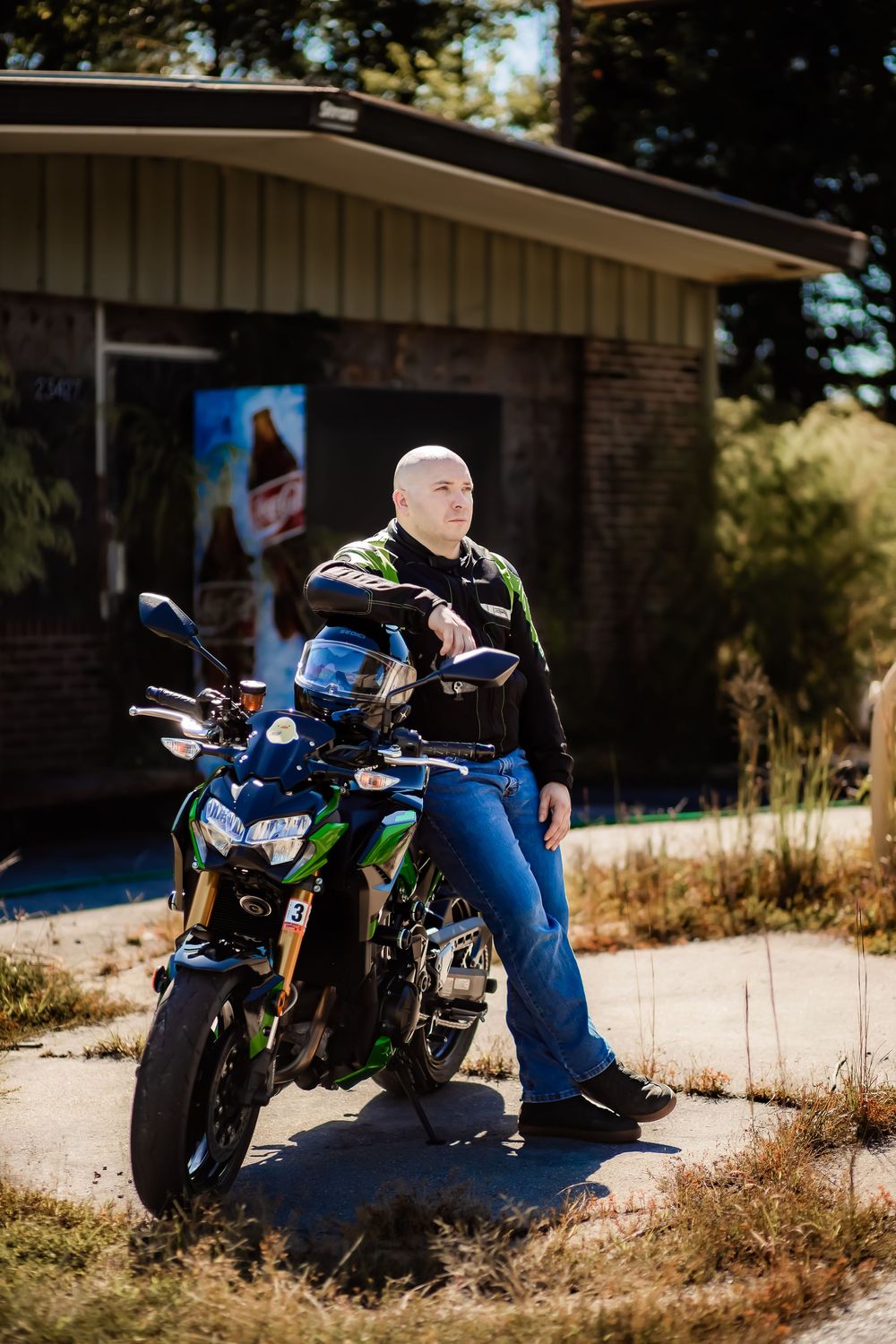 Bald man in motorcycle gear leaning on a green Kawasaki sport bike outside an abandoned building.