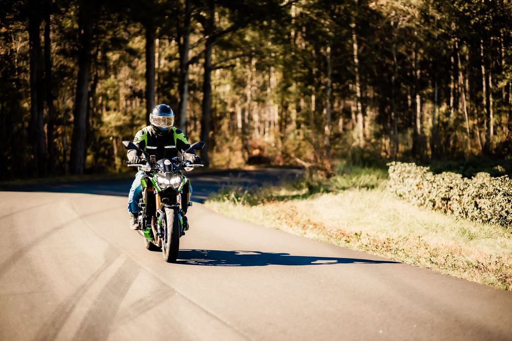 Motorcyclist riding a green bike on a winding forest road wearing a helmet and protective gear.