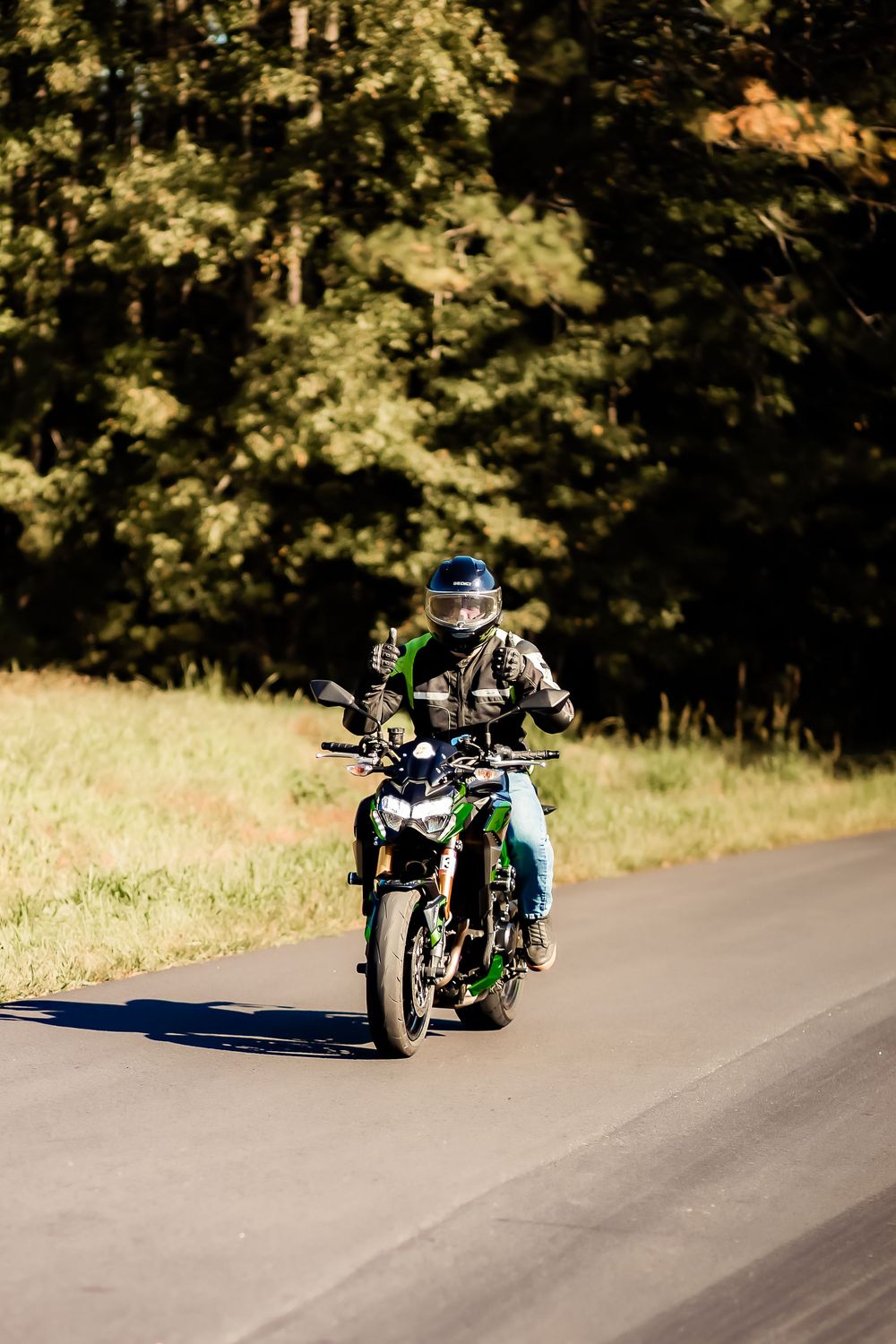 Motorcyclist riding a green and black bike on a rural road, wearing a helmet and camo jacket, giving a thumbs up.