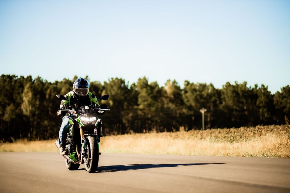 Motorcyclist in green gear riding a black motorcycle on an open road with trees in the background on a clear day.