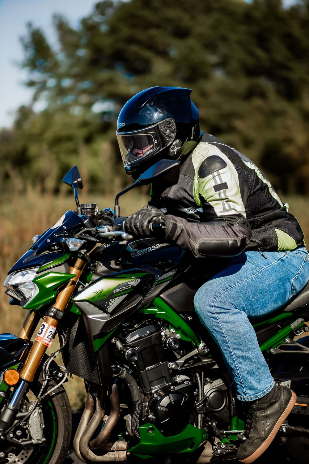A motorcyclist in a black helmet and jacket rides a green Kawasaki motorcycle on a rural road.