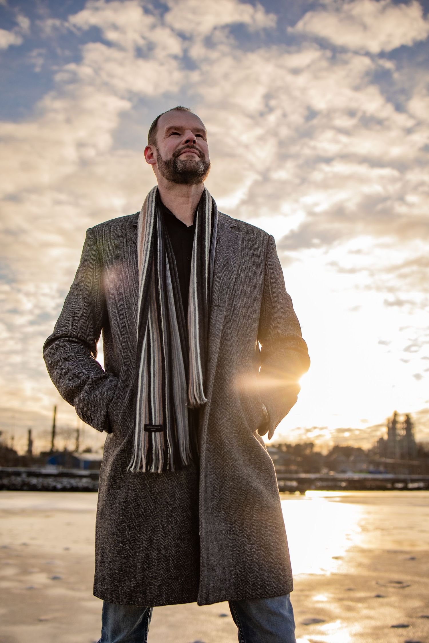 Man in gray wool coat and striped scarf standing outdoors at sunset with dramatic cloudy sky backdrop.