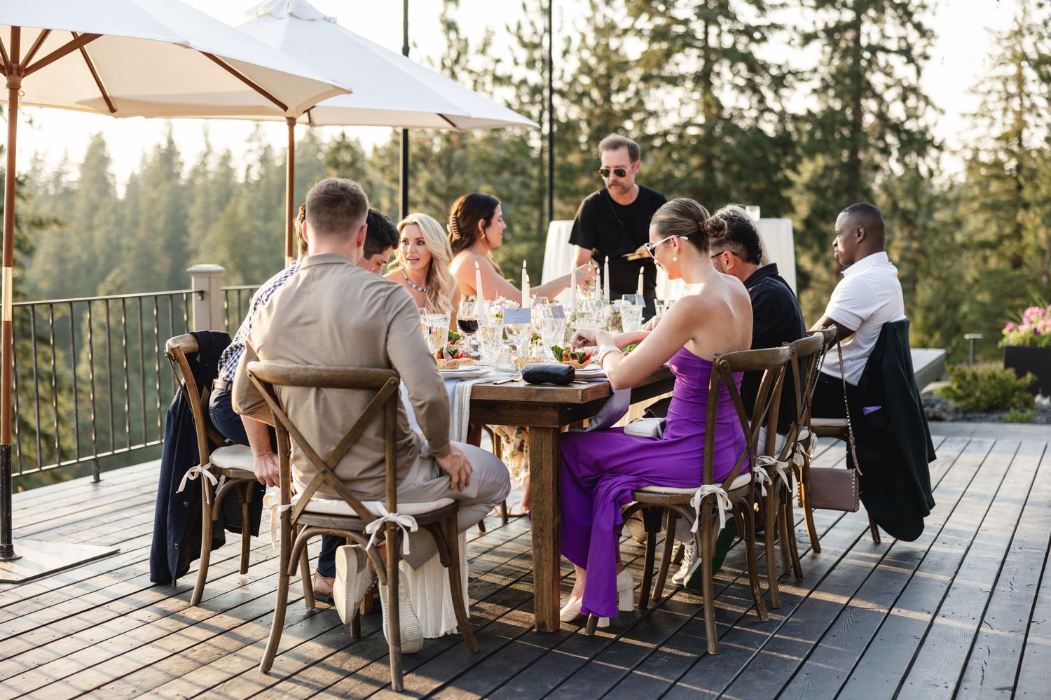 Group of friends dining al fresco on a wooden deck surrounded by tall pine trees at golden hour.
